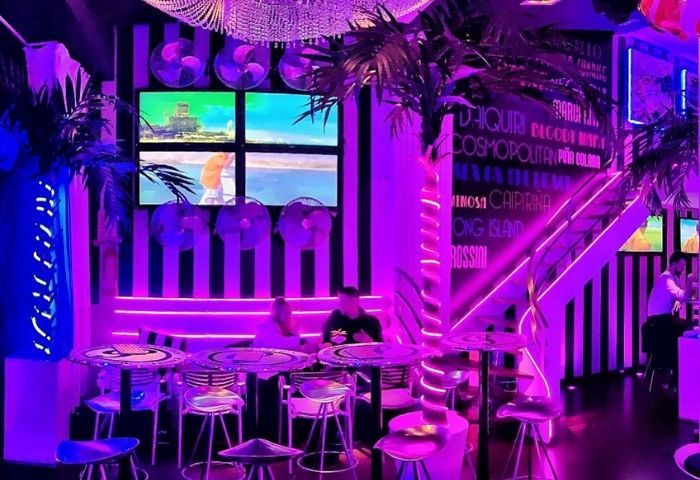 Barcelona's Plata Cocktail Bar, a gay bar, glows purple. Palm trees, fans, & screens add flair. Tables & chairs await patrons.