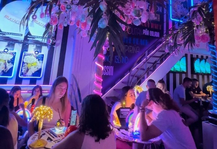 Barcelona's Plata Cocktail Bar, a gay bar, has a vibrant interior. People sit at tables enjoying drinks under neon lights and palm tree decor.