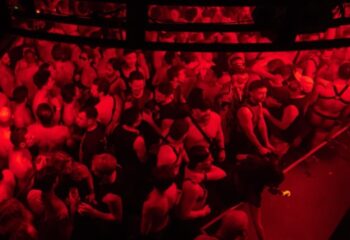 Red-lit crowd at Poof Doof, a gay dance party in Melbourne, Australia. Men in harnesses dance. Gay travel advice.