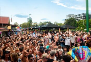 Crowd at Poof Doof Melbourne, a gay dance party. People dancing, flags, fans, and black umbrellas. Outdoor event, travel advice.