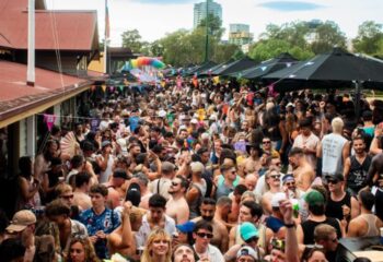 Crowd at Poof Doof Melbourne, a gay party, with rainbow decorations and flags. People are dancing and enjoying the outdoor event.