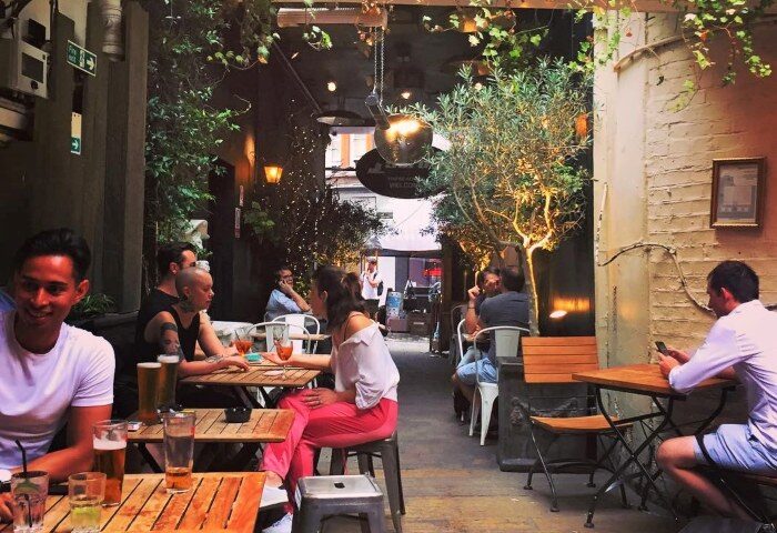 Outdoor seating area at The Yard Bar, a popular gay bar in Soho, London. People are enjoying drinks at tables surrounded by greenery in the city centre.