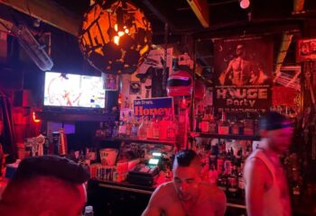 Inside Powerhouse Gay Bar in San Francisco. A bartender stands in front of shelves of liquor. A TV plays over the bar. Red lighting.