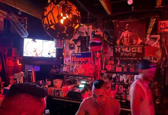 Inside Powerhouse Gay Bar in San Francisco. A bartender stands in front of shelves of liquor. A TV plays over the bar. Red lighting.