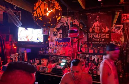 Inside Powerhouse Gay Bar in San Francisco. A bartender stands in front of shelves of liquor. A TV plays over the bar. Red lighting.