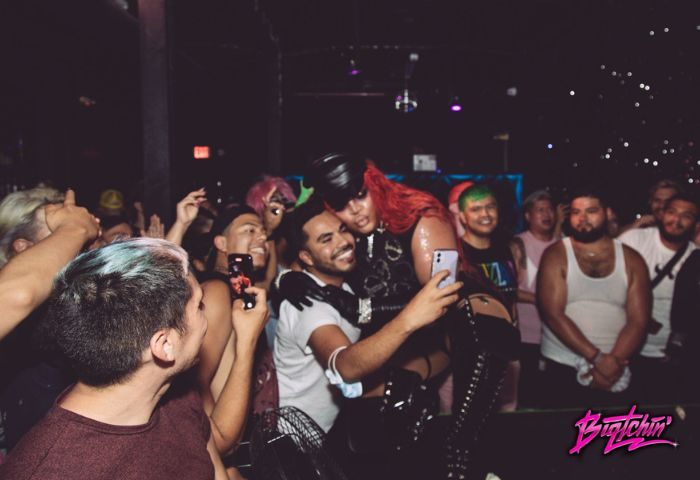 Precinct Party: Gay men at a party take a selfie with a drag queen in fetish gear. People are holding up their phones to take pictures.