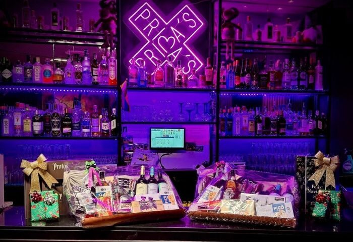 Prima's S-Bar, a Gay Bar in Madrid. Shelves of liquor bottles are lit with purple neon. Gift baskets sit on the bar. Madrid gay nightlife.