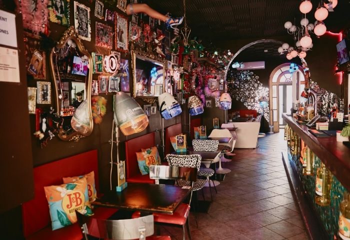 Interior of Priscilla Cafe, a gay bar in Barcelona. Eclectic decor with photos, mirrors, vintage hair dryers, and a bar area. Gay travel.