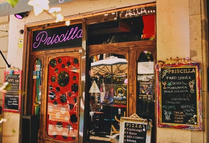 Priscilla Cafe-Bar, Barcelona. Wooden door with neon sign. Tapas & cocktail menu boards. Gay travel advice for best gay bars.