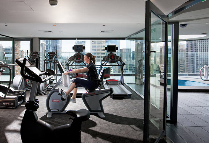 Pullman Sydney Hyde Park gym. Woman using exercise bike. Rooftop pool view. Gay hotel in Darlinghurst, Sydney. Affordable luxury.