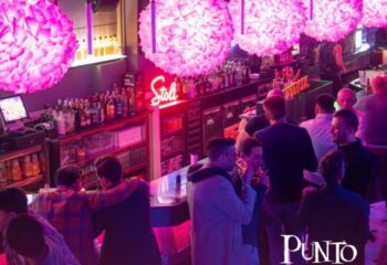 Inside Punto BCN, a gay bar in Barcelona, with pink lighting, floral lamps, and men socializing at the bar. Nightlife scene.
