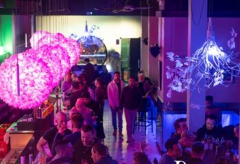 Interior view of Punto BCN Gay Bar in Barcelona. Crowded space with pink lighting, bar areas, and people socializing. Gay travel destination.