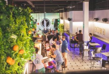 Interior shot of Punto BCN, a gay bar in Barcelona. Men are socializing at the bar and tables. Green wall with oranges visible.