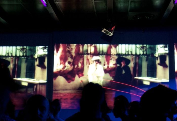 QClub Milan: Gay club interior with a video art installation on a screen, featuring figures in hats and abstract imagery. Nightlife scene.