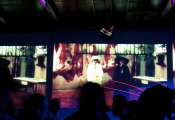QClub Milan: Gay club interior with a video art installation on a screen, featuring figures in hats and abstract imagery. Nightlife scene.