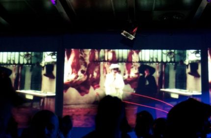 QClub Milan: Gay club interior with a video art installation on a screen, featuring figures in hats and abstract imagery. Nightlife scene.