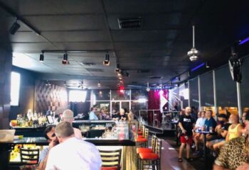 QUADZ Experience: Interior of a gay bar, with men socializing at the bar and tables. Stage with rainbow flag. Gay travel advice.