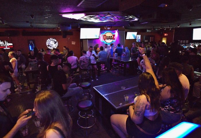 Inside Quadz Video Bar, Las Vegas. A lively gay bar scene with patrons at tables, stools, and the bar. TVs and colorful lighting add to the ambiance.
