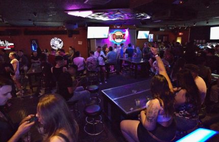 Inside Quadz Video Bar, Las Vegas. A lively gay bar scene with patrons at tables, stools, and the bar. TVs and colorful lighting add to the ambiance.
