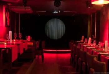 Interior of Queenz Cabaret Show gay bar in Sitges. Tables set for a show, red lighting, stage with a curtain. Gay travel destination.