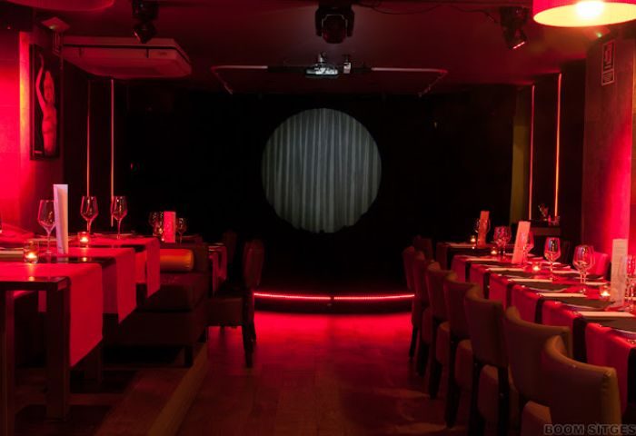 Interior of Queenz Cabaret Show gay bar in Sitges. Tables set for a show, red lighting, stage with a curtain. Gay travel destination.