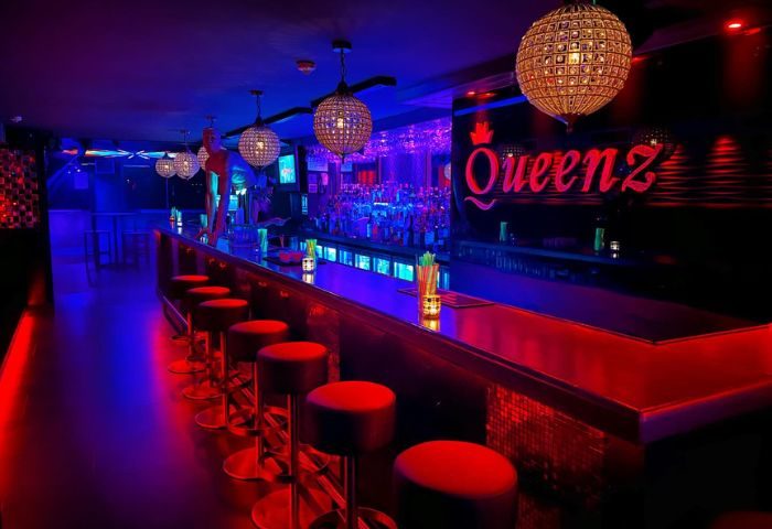 Queenz Music Bar, a gay club in Sitges. Red bar stools line the counter under disco ball lights. Bottles line the bar. Nightlife scene.