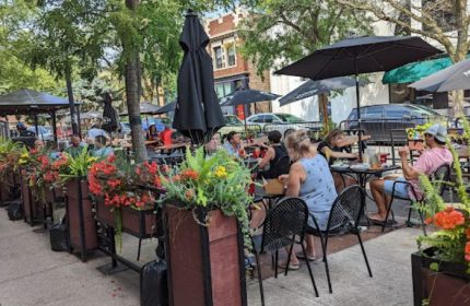 Outdoor seating at REPLAY Andersonville, a gay bar with food in Chicago. People are enjoying drinks under umbrellas on a sunny day.