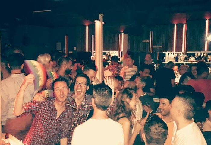Gay bar in Madrid: crowded scene of men socializing, some holding rainbow flags. Gay travel advice for nightlife in Spain.