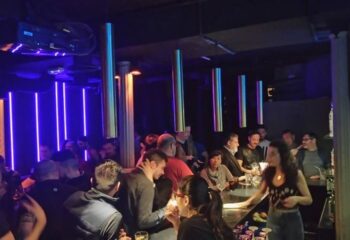 Rick's Gay Bar in Madrid. Interior view of the bar with people gathered around the counter, illuminated by blue neon lights. Gay travel advice.