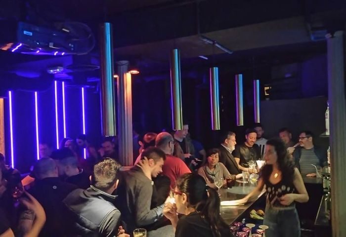 Rick's Gay Bar in Madrid. Interior view of the bar with people gathered around the counter, illuminated by blue neon lights. Gay travel advice.