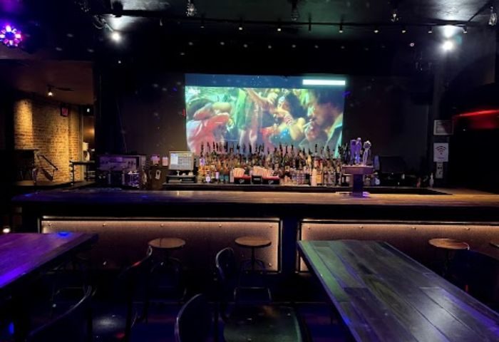 Rebar Chelsea in New York: Interior shot of the gay bar. A large screen shows a crowd, bar stools line the counter, tables in the foreground.