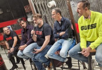 Group of men at Rectum Gay Cruise Club in Barcelona. Men sit on stools, one speaks into a microphone. Gay travel advice.