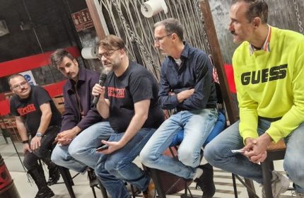 Group of men at Rectum Gay Cruise Club in Barcelona. Men sit on stools, one speaks into a microphone. Gay travel advice.