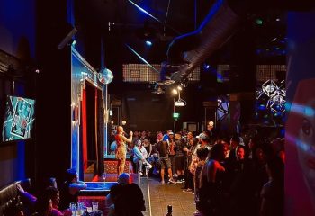 Redline Shows: Gay bar scene with performer on stage. Crowd of gay men enjoying the show. Nightlife in a gay-friendly venue. Travel advice.