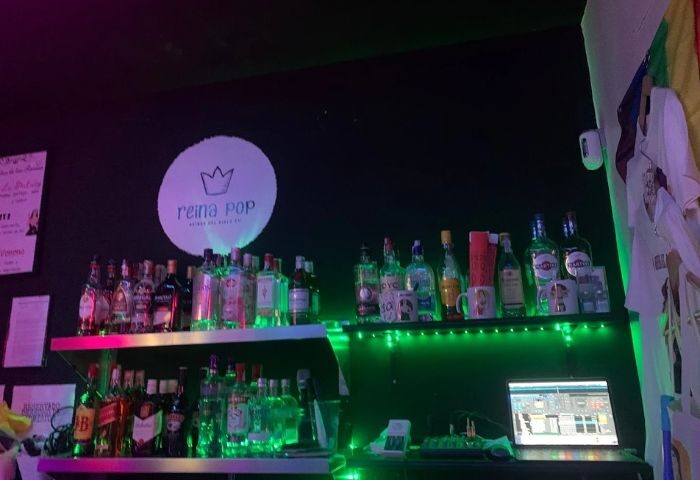 Reina Pop Gay Bar in Madrid: Shelves stocked with liquor bottles and a laptop, illuminated by green neon lights in a vibrant gay nightlife scene.