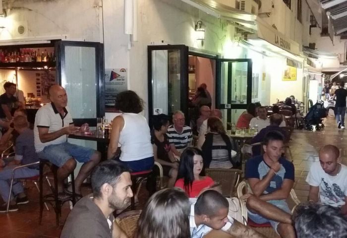 Patrons at René's Bar La Esquina, a gay bar in Torremolinos, Spain. Outdoor seating area with people enjoying drinks and conversation.