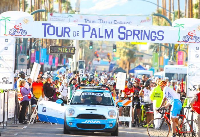 Tour de Palm Springs event. Cyclists line up with a pace car at the start line. A banner reads Tour de Palm Springs. Nearby events for gay men.