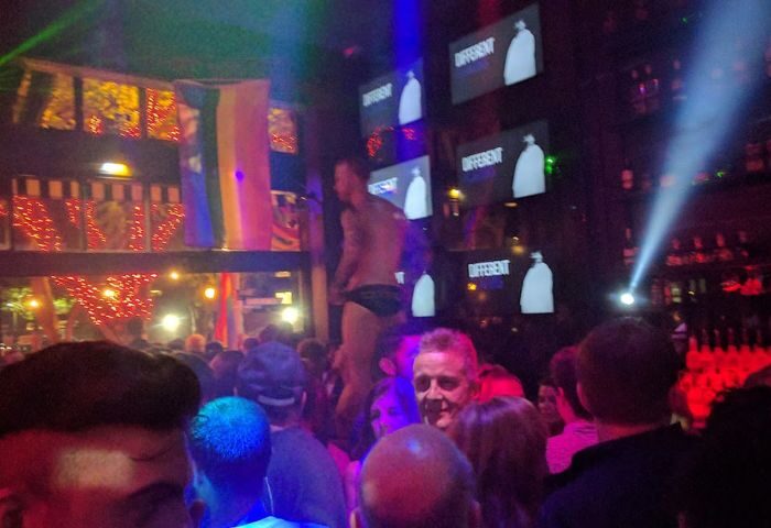 Gay Revolver party scene: man in underwear stands above crowd in a nightclub with rainbow flag. Gay men travel advice.