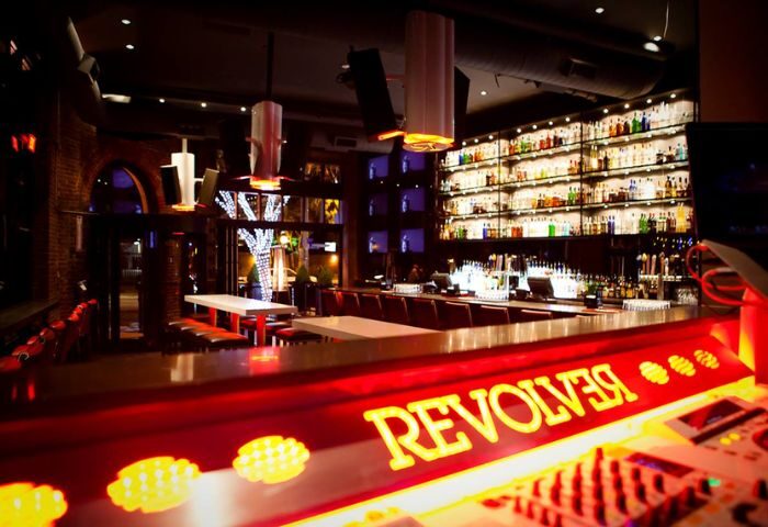 Interior of Revolver, a gay bar. Lit bar with liquor shelves, tables, and DJ equipment. Gay travel nightlife spot.