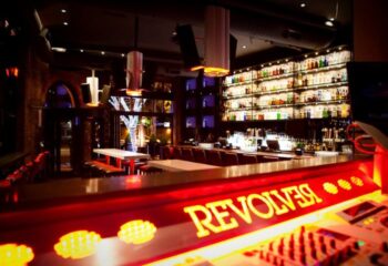 Interior of Revolver, a gay bar. Lit bar with liquor shelves, tables, and DJ equipment. Gay travel nightlife spot.
