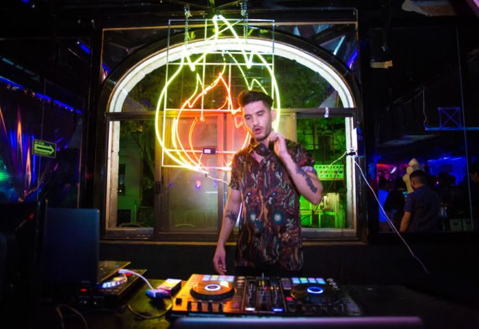 DJ at Rico Club, a gay bar in Mexico. Man with tattoos and headphones stands behind DJ equipment, neon lights glow in background.