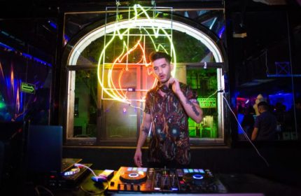 DJ at Rico Club, a gay bar in Mexico. Man with tattoos and headphones stands behind DJ equipment, neon lights glow in background.