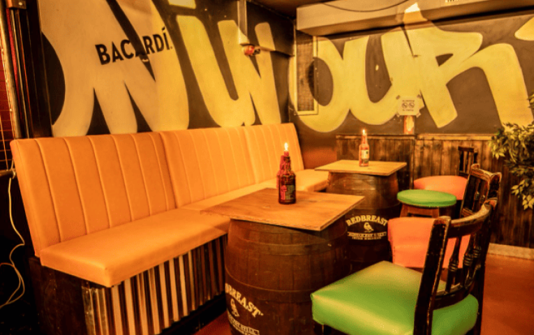 Riot Gay Bar Dublin interior. Cozy seating area with orange bench, barrel tables, and colorful chairs. Graffiti art on the walls. Gay travel.
