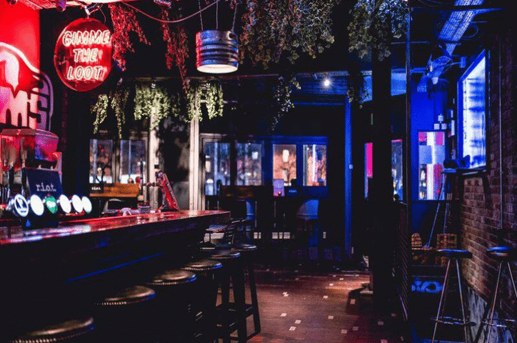 Interior of Riot Gay Bar in Dublin. Bar with stools, neon sign reading Gimme the Loot, plants hanging from the ceiling, and dim lighting.