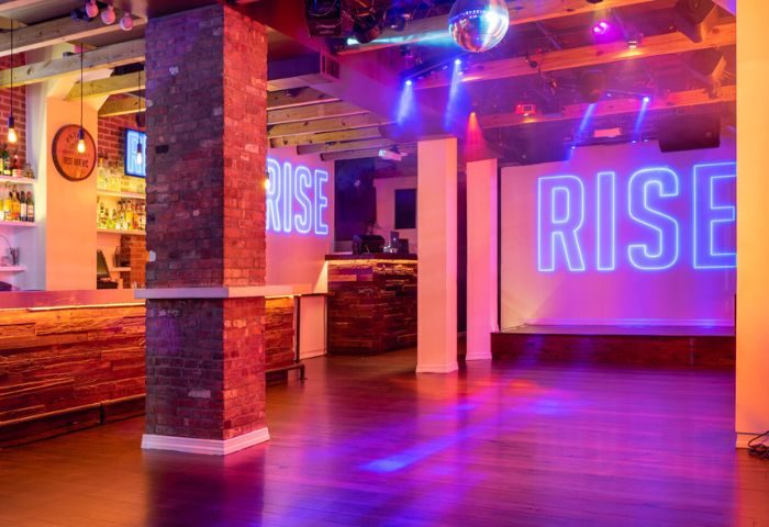 Interior of Rise Gay Bar in New York. The dance floor is lit with purple and blue lights, and the bar is stocked with drinks. Disco ball.