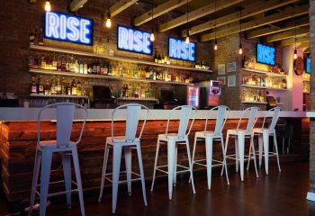 Rise Bar in New York: Interior view of the gay bar featuring white stools at the bar, neon signs, and a well-stocked liquor shelf.