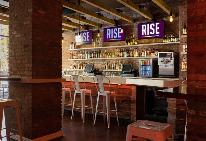 Rise Gay Bar in New York: Interior view of the bar area with exposed brick, wooden beams, liquor shelves, and screens displaying RISE.