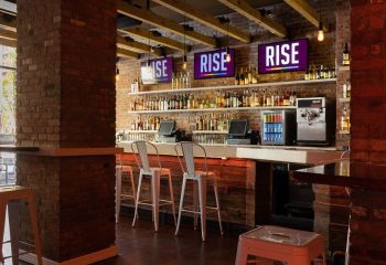 Rise Gay Bar in New York: Interior view of the bar area with exposed brick, wooden beams, liquor shelves, and screens displaying RISE.