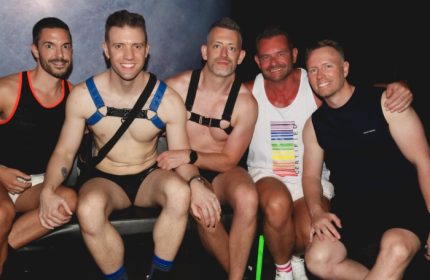 Five attractive gay men pose at a Rock City party in Melbourne. Some wear harnesses, tank tops, and briefs. Nightlife and gay travel.