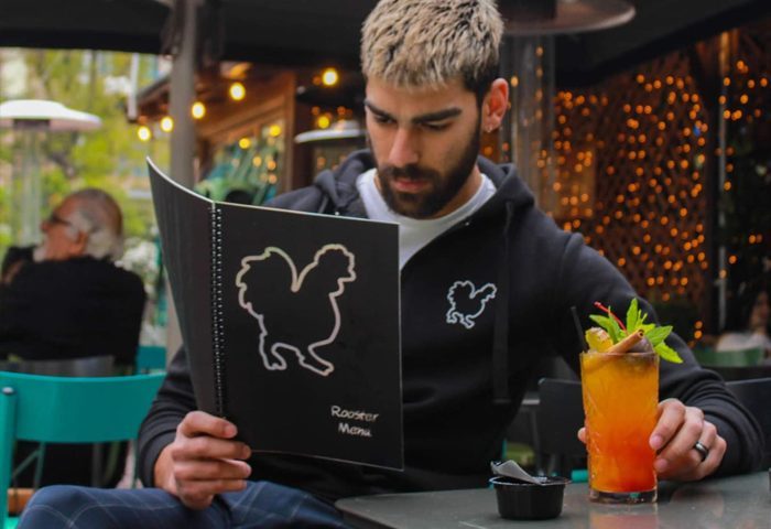 Gay man at Rooster Athens bar, reading the menu. Cocktail on the table. Rooster logo on menu and hoodie. Gay travel advice.